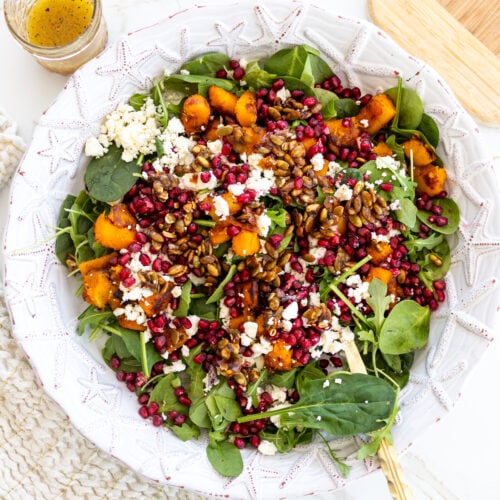 A vibrant butternut squash and arugula salad sits in a white bowl, showcasing roasted pumpkin chunks, pomegranate seeds, crumbled feta cheese, and spiced pumpkin seeds. A jar of vinaigrette accompanies it on a white table with a woven placemat.