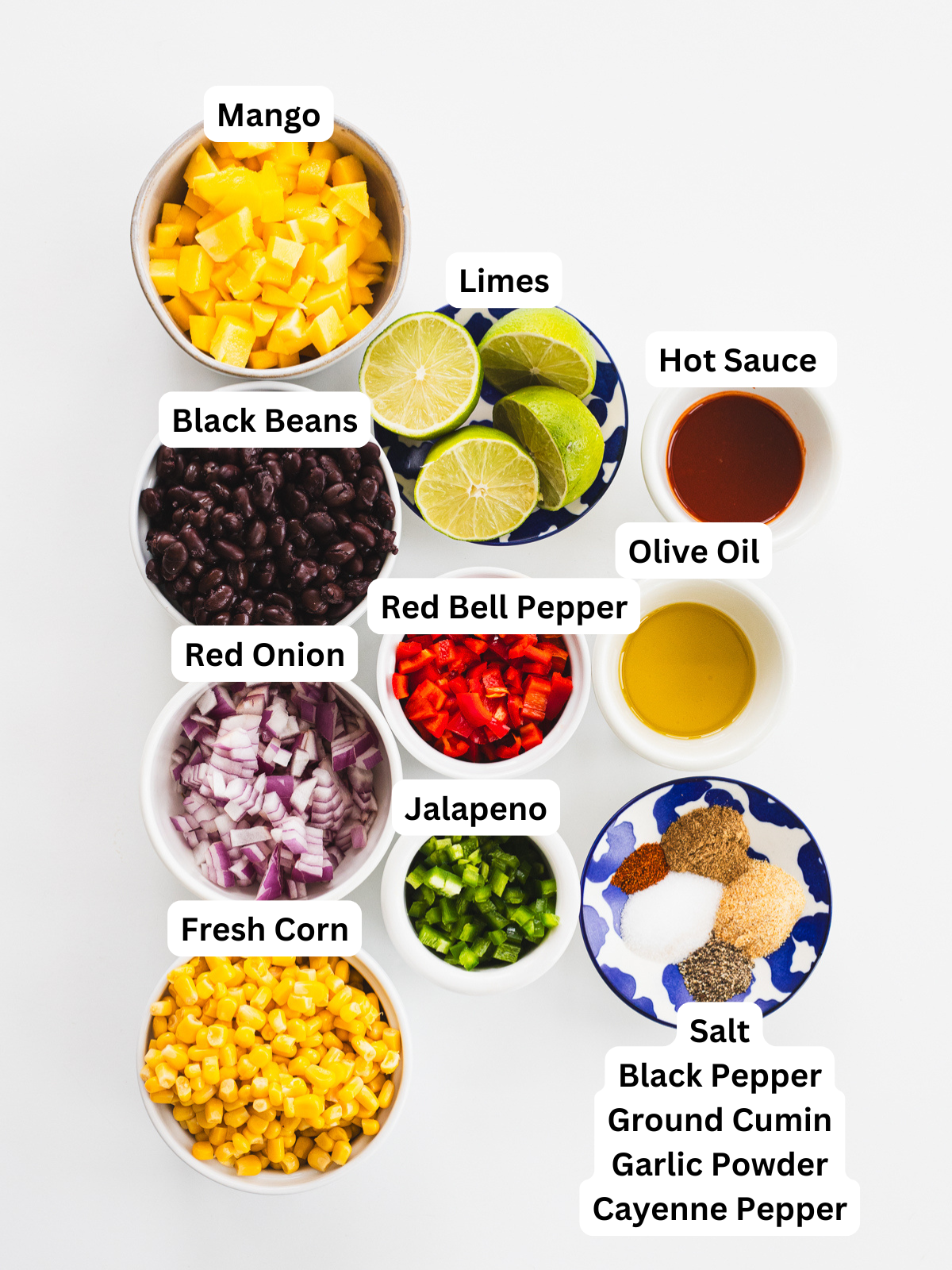 Overhead view of bowls with labeled ingredients for black bean corn salad: diced mango, limes, black beans, hot sauce, olive oil, red bell pepper, red onion, jalapeño, fresh corn, and spices (salt, black pepper, cumin, garlic powder, cayenne).
