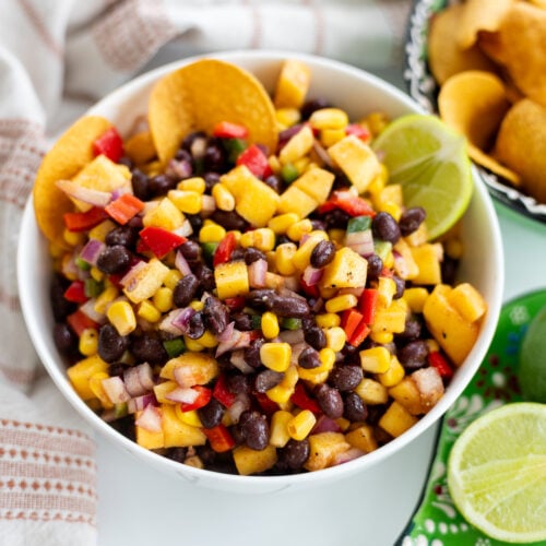 A vibrant black bean corn salad salsa with diced red peppers, red onions, and mango, garnished with lime wedges and served alongside crispy round tortilla chips.