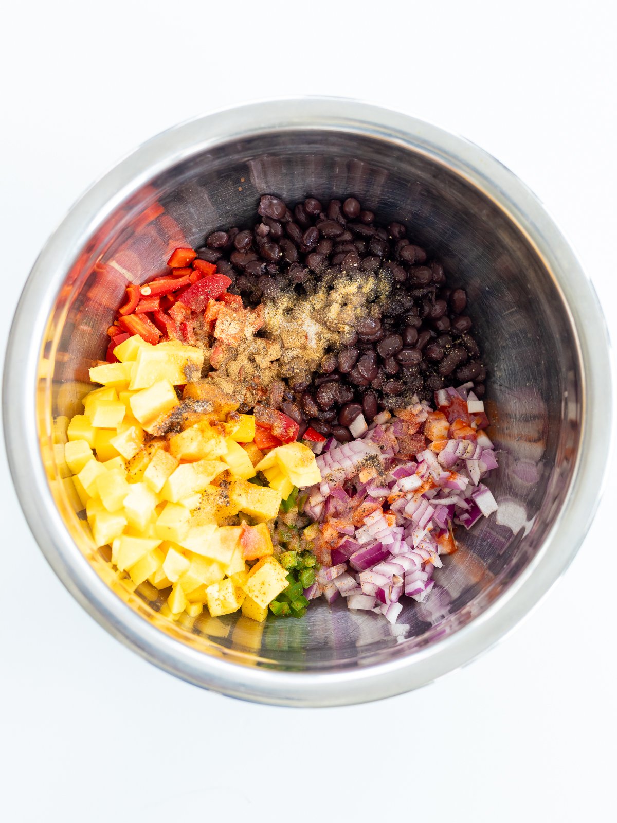 A metal mixing bowl filled with chopped mango, corn, red bell peppers, red onions, black beans, and spices—perfect ingredients for a colorful black bean corn salad—arranged in separate sections on a white background.