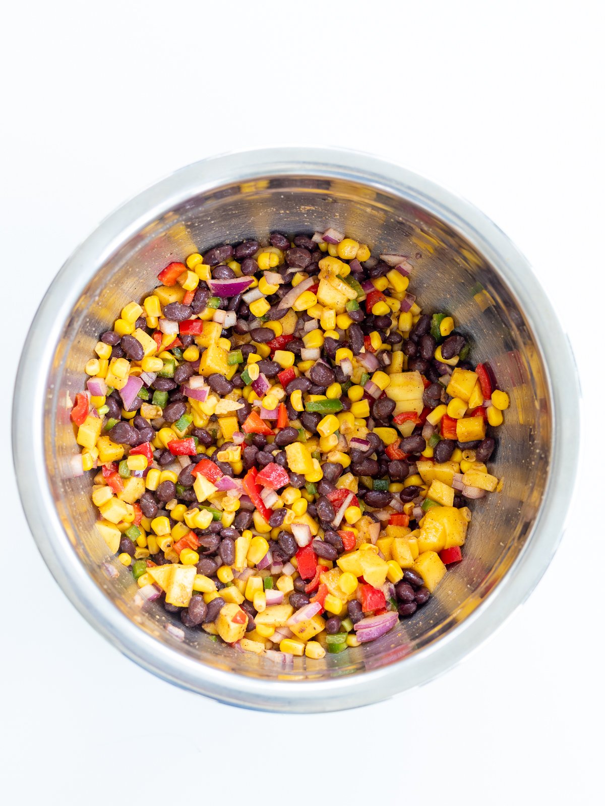 A stainless steel bowl filled with a colorful black bean corn salad, featuring diced red onions, red bell peppers, fresh corn, and chopped mango on a white background.