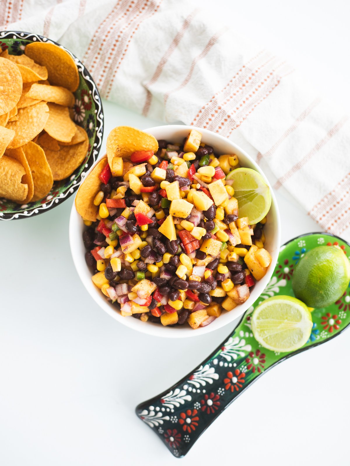 A bowl of black bean corn salad with red pepper salsa, lime wedges, and Fritos chips on the side, next to a bowl of chips and a decorative spoon holding two halved limes on a white background.