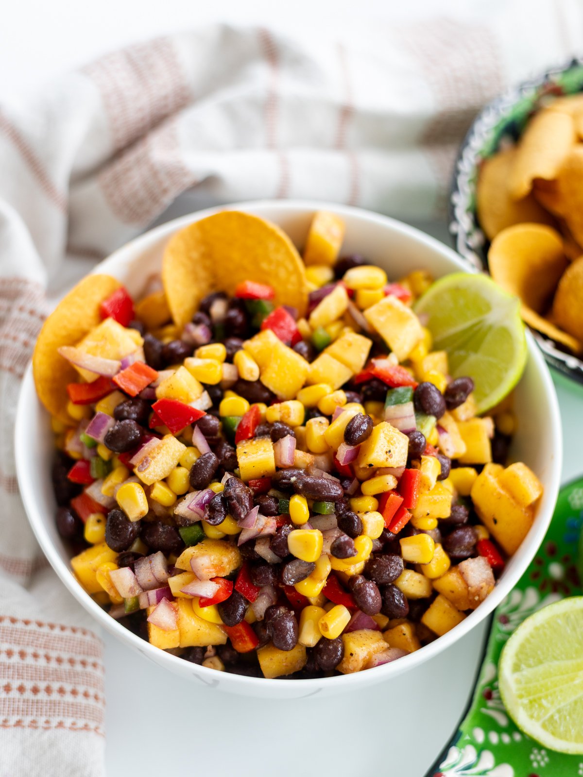 A bowl of black bean corn salad with diced red peppers, onions, and mango is garnished with lime wedges and served with tortilla chips. A patterned plate and white towel are visible in the background.
