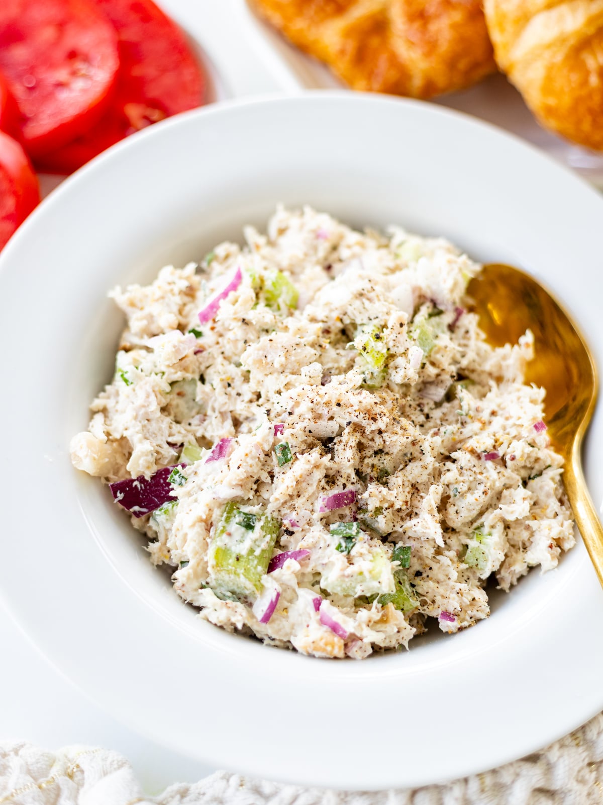 A white bowl filled with creamy turkey salad containing chopped celery, red onion, and herbs, topped with black pepper. A gold spoon rests on the side, with sliced tomatoes and croissants in the background.