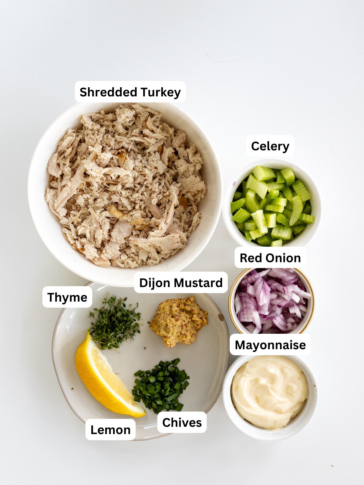 A bowl of shredded turkey, and small bowls of chopped celery, red onion, mayonnaise, Dijon mustard, chopped thyme, chopped chives, and a lemon wedge, all labeled, arranged on a white background.