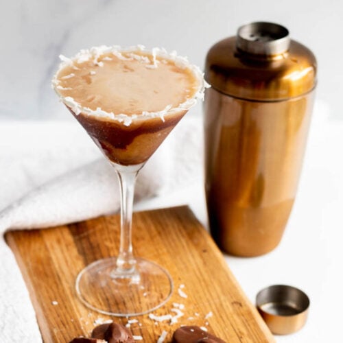 An Almond Joy Martini with a cocktail shaker and Almond Joy candy bars on a wooden tray.