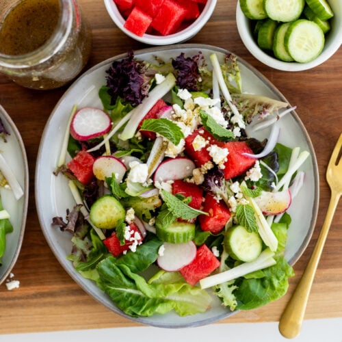 Watermelon salad on a plate with extra dressing, watermelon chunks and cucumber slices in bowls.