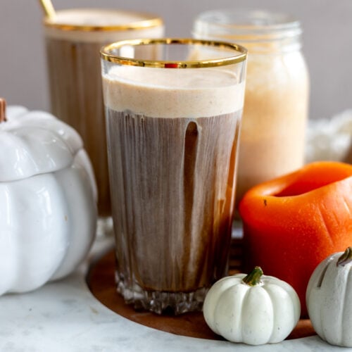 A frothy iced coffee drink in a gold-rimmed glass sits on a wooden tray adorned with small white and orange pumpkins and a large orange candle. In the background is another glass of the same drink, and a jar filled with an orange-colored beverage.