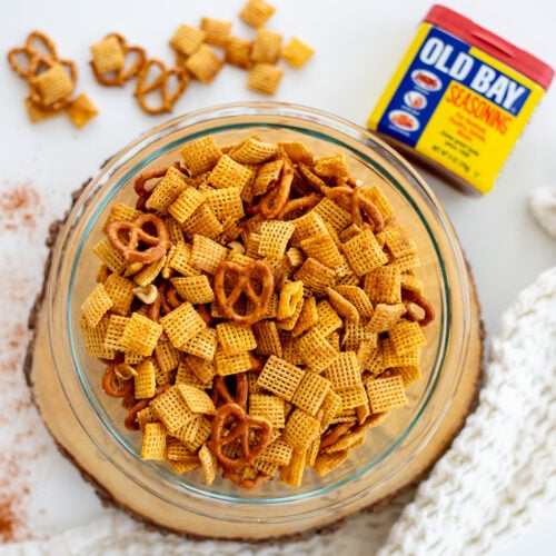 A glass bowl filled with a savory Old Bay Chex Mix, featuring square cereal pieces and pretzels, seasoned to perfection. The mix is placed on a wooden surface, with additional snack pieces and a can of Old Bay seasoning visible in the background.