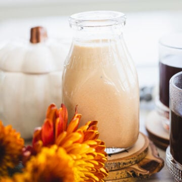 A glass bottle filled with a creamy orange pumpkin spice coffee creamer is surrounded by autumn decor, including vibrant orange chrysanthemums, a small white pumpkin, and a glass of black coffee on wooden coasters. Coffee beans are scattered nearby.