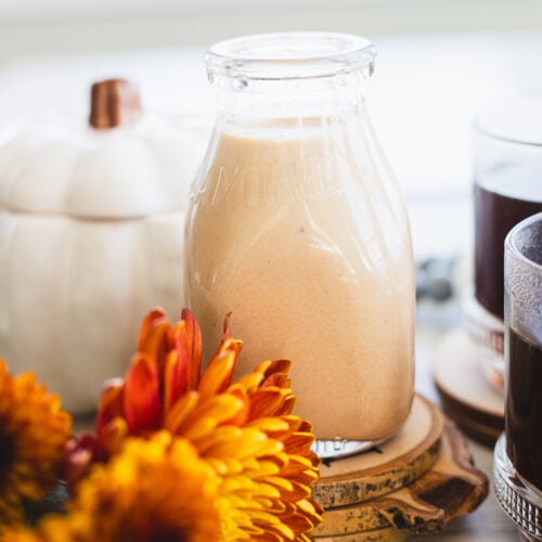 A glass bottle filled with a creamy orange pumpkin spice coffee creamer is surrounded by autumn decor, including vibrant orange chrysanthemums, a small white pumpkin, and a glass of black coffee on wooden coasters. Coffee beans are scattered nearby.
