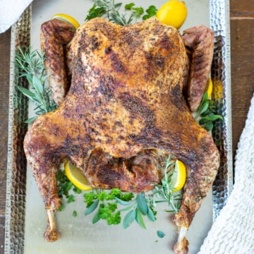 A roasted turkey on a tray, surrounded by lemon slices and fresh herbs like parsley and rosemary, garnished for presentation. The turkey is golden-brown and placed on a textured metal serving dish.