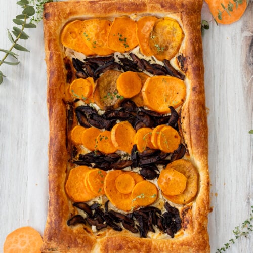 A rectangular tart with golden brown pastry, topped with sliced sweet potatoes and mushrooms. Garnished with herbs. Surrounding the tart are sprigs of greenery and an orange vegetable slice on a light wooden surface.
