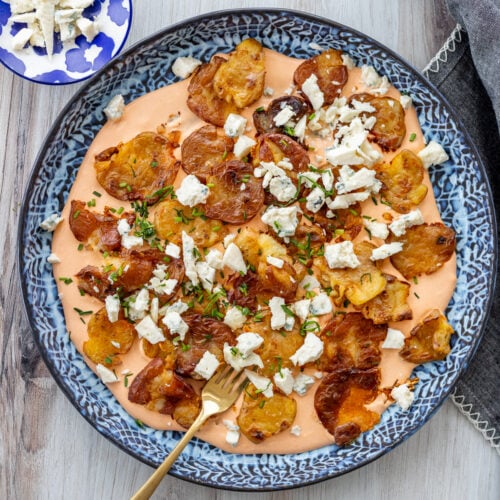 A plate of crispy smashed potatoes topped with crumbled cheese and chopped herbs, served on a buffalo sauce. There's a gold fork on the side. A small dish with more cheese crumbles is partially visible in the background.
