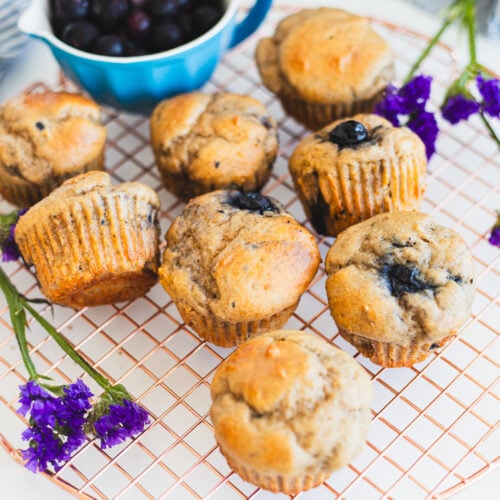 Seven blueberry banana muffins on a wire cooling rack, surrounded by purple flowers. A small blue bowl filled with fresh blueberries is in the background. The golden-brown muffins are studded with visible blueberries, creating a delicious fusion of flavors and textures.