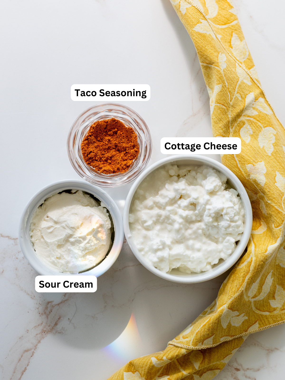 Three bowls with labeled ingredients on a white surface: taco seasoning in a small glass bowl, cottage cheese for Cottage Cheese Taco Dip in a larger bowl, and sour cream in another; a yellow patterned cloth lies beside them.