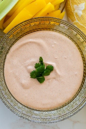 A glass bowl of creamy Cottage Cheese Taco Dip garnished with cilantro, surrounded by fresh vegetable sticks and a yellow patterned napkin.