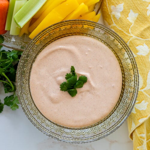 A glass bowl of creamy Cottage Cheese Taco Dip garnished with cilantro, surrounded by fresh vegetable sticks and a yellow patterned napkin.