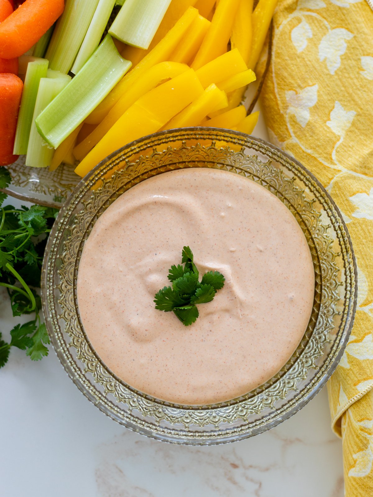 A glass bowl filled with creamy, light orange Cottage Cheese Taco Dip garnished with cilantro, surrounded by cut celery, carrots, yellow bell pepper, and a yellow patterned napkin.