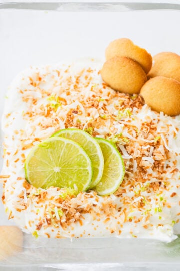 A glass dish with creamy key lime pie dip, topped with toasted coconut flakes, three fresh lime slices, and three vanilla wafer cookies on one side.