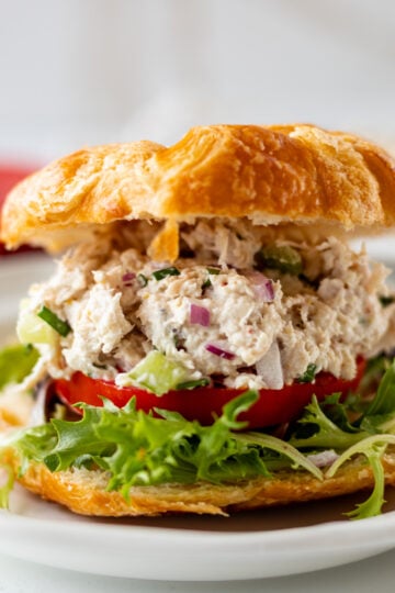 A croissant sandwich filled with leafy greens, tomato slices, and a creamy turkey salad with chopped vegetables, served on a white plate.