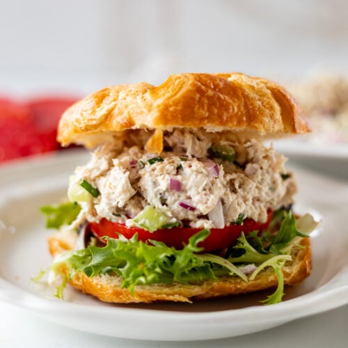 A croissant sandwich filled with leafy greens, tomato slices, and a creamy turkey salad with chopped vegetables, served on a white plate.